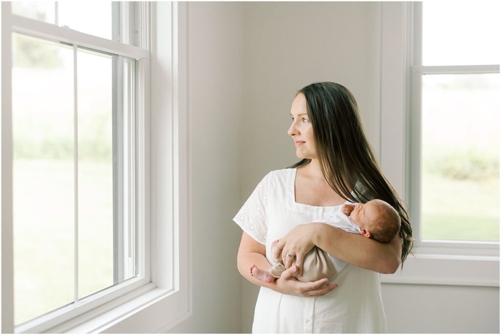 Mother looking out the window holding her newborn with Angelique Jasmin Photography