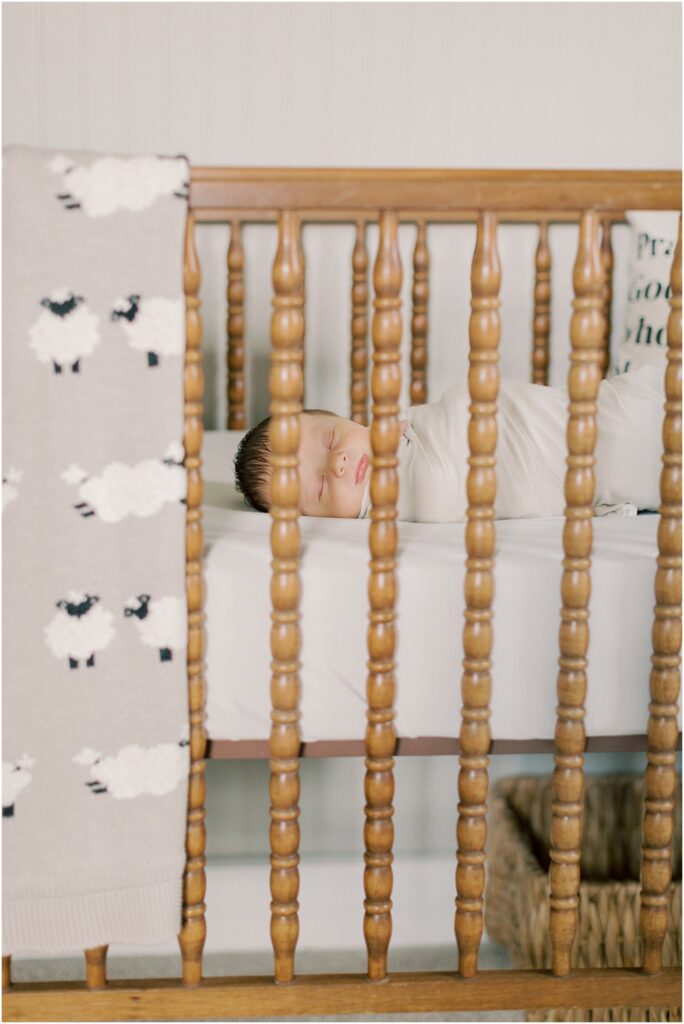 Newborn sleeping in his crib with a sheep blanket hanging on the side of the crib in a lifestyle newborn photography session.