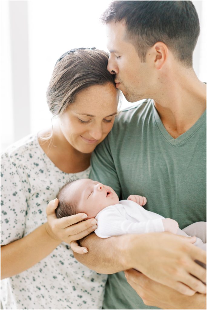 Dad kissing mama at newborn session with baby in his arms at summer indoor newborn session