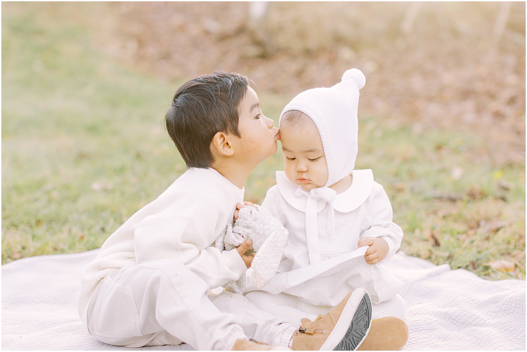Young toddler boy kissing baby sister while sitting on a blanket