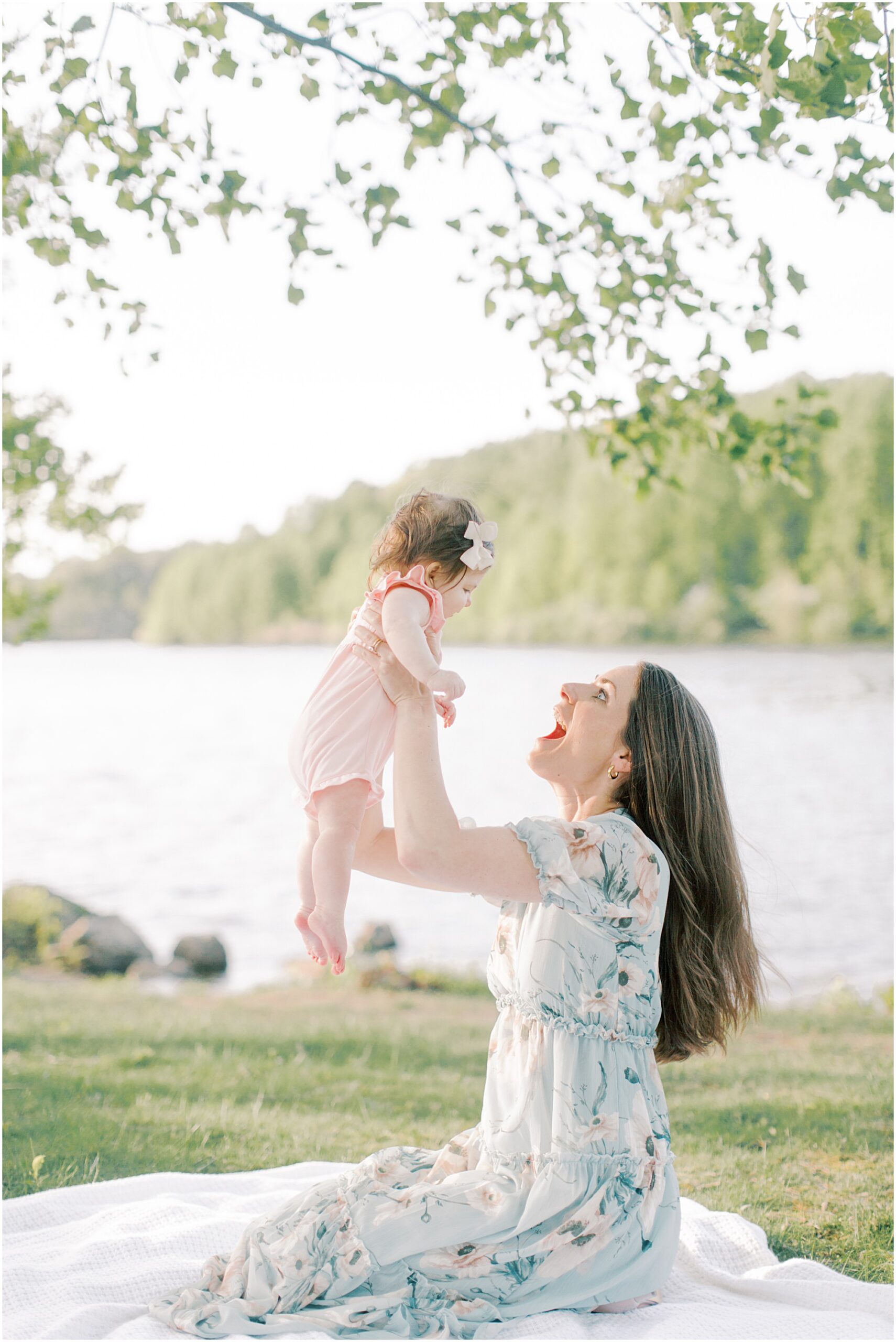 Mother holding up baby girl in the air while sitting on a blanket next to the lake
