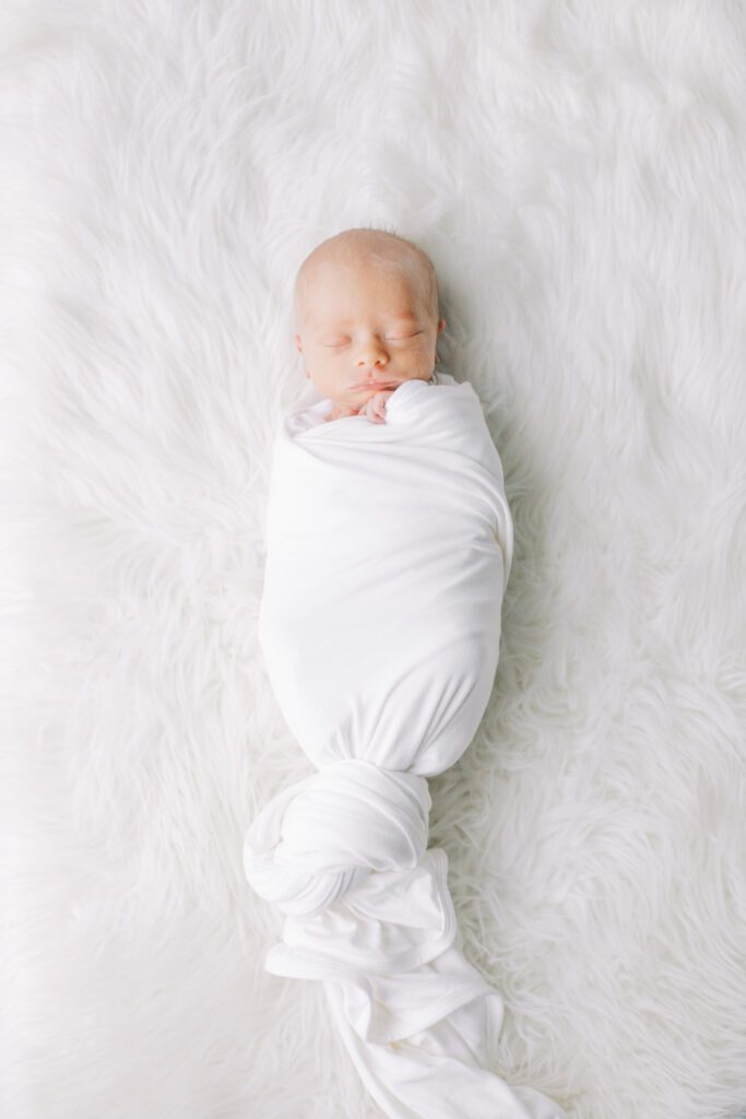 Newborn baby wrapped in white swaddle blanket on white fur background