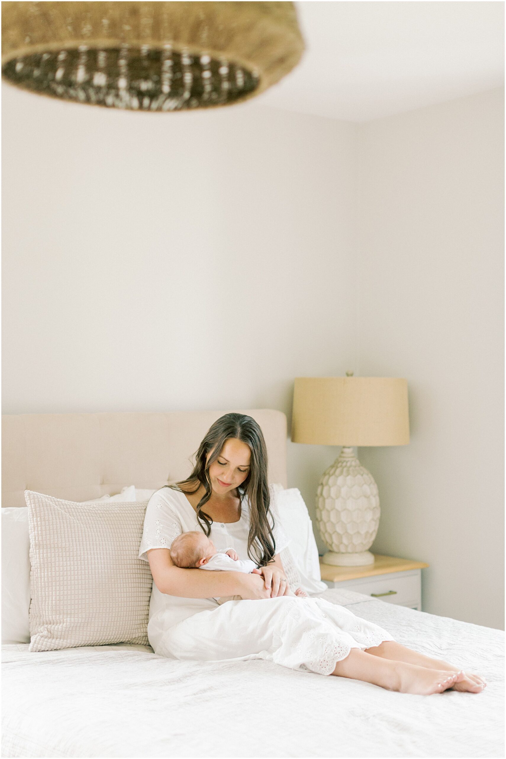 Mama sitting on her white bed holding her newborn son with Chester Count Pa Newborn Photographer Angelique Jasmin Photography