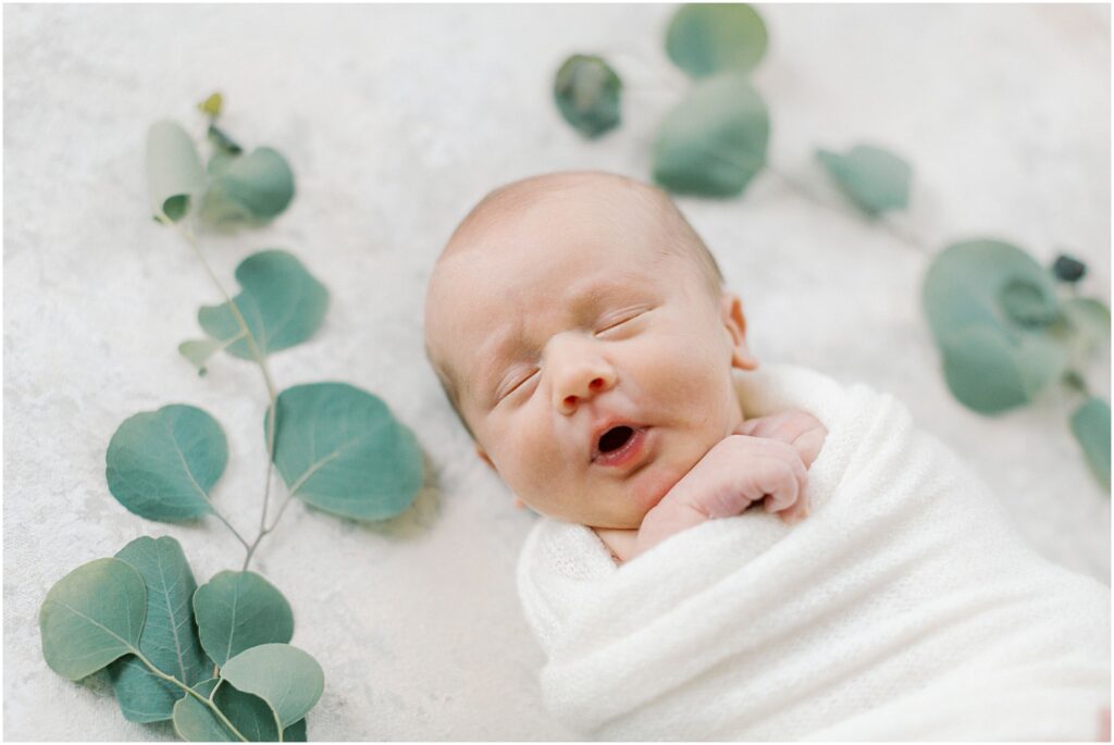 Newborn baby boy with mouth in an open "o" on a styling mat with eucalyptus. 