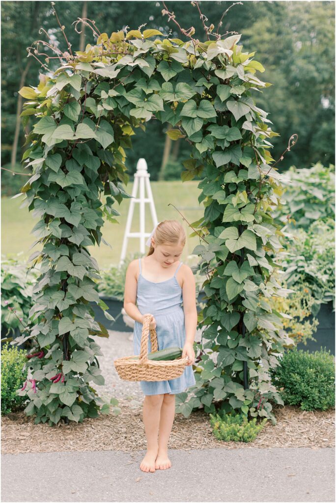 Young girl standing in front of a garden arbor with a basket of produce.