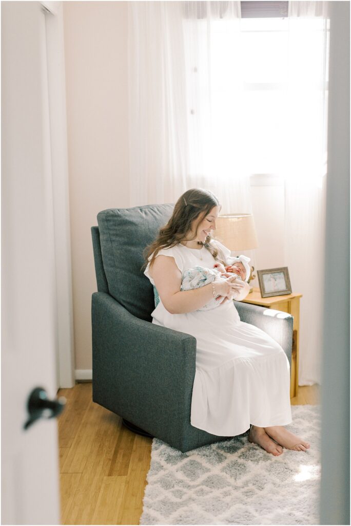 looking through a doorway at a mother sitting in a chair holding her newborn baby girl in Elizabethtown PA Newborn Photographer.