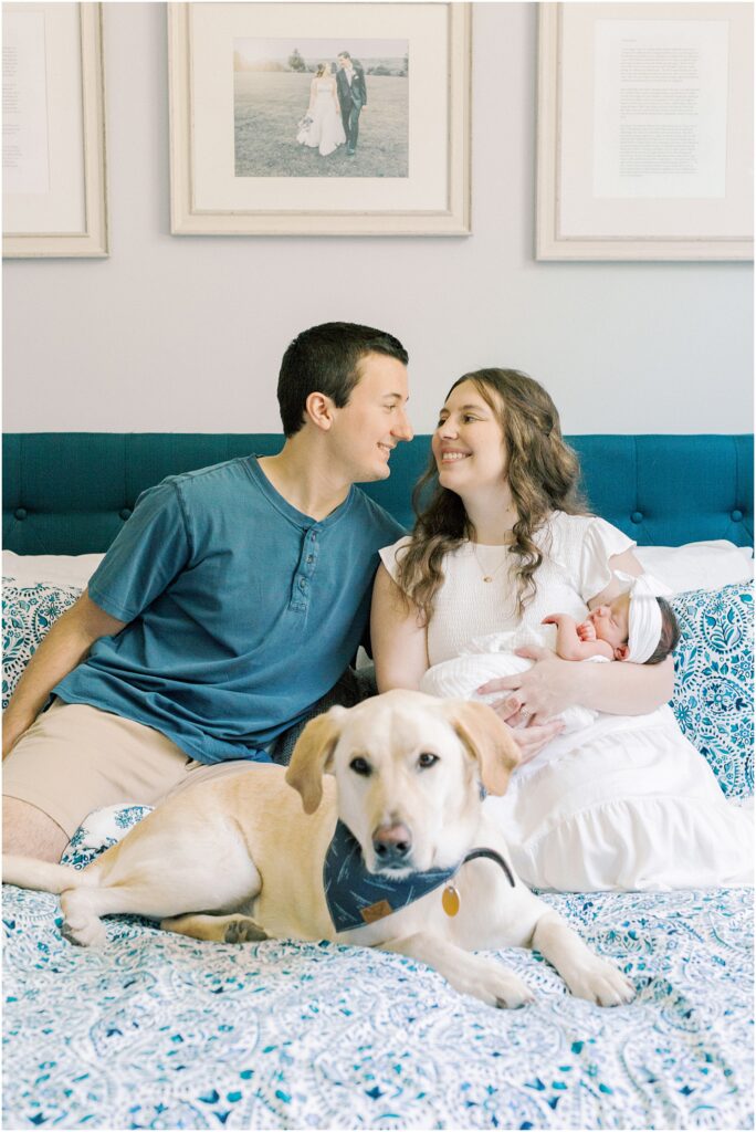 Parent's sitting on the bed looking at each other with their newborn baby in their arms with a dog in the foreground.