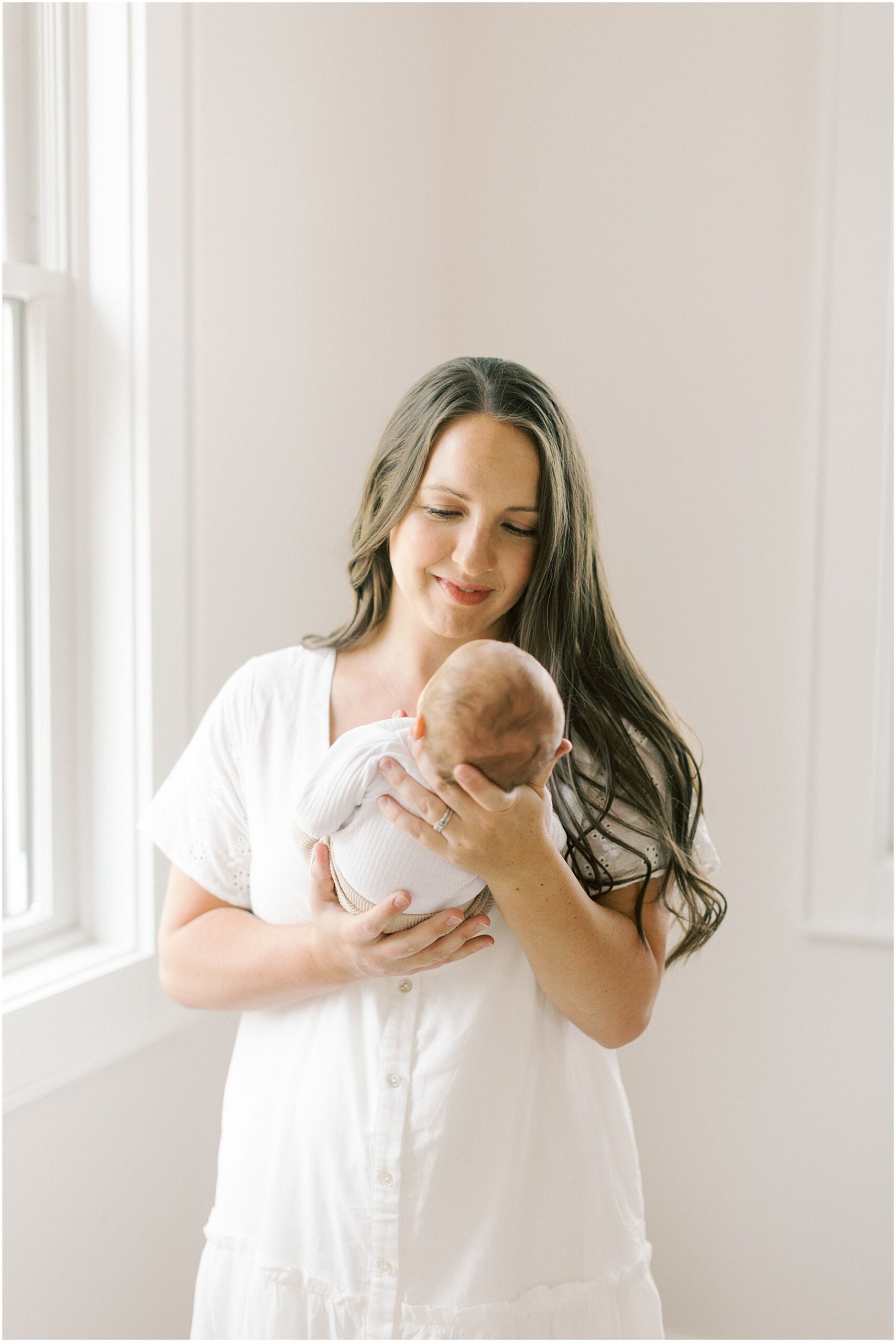 Mother in white dress standing near a window looking at her newborn son in when to book newborn photos.