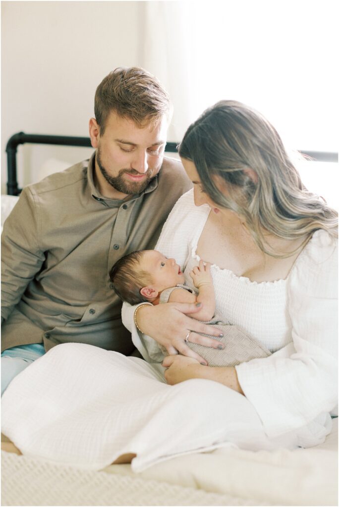 Father and Mother looking down on their baby boy while sitting on their bed for their lifestyle newborn session.