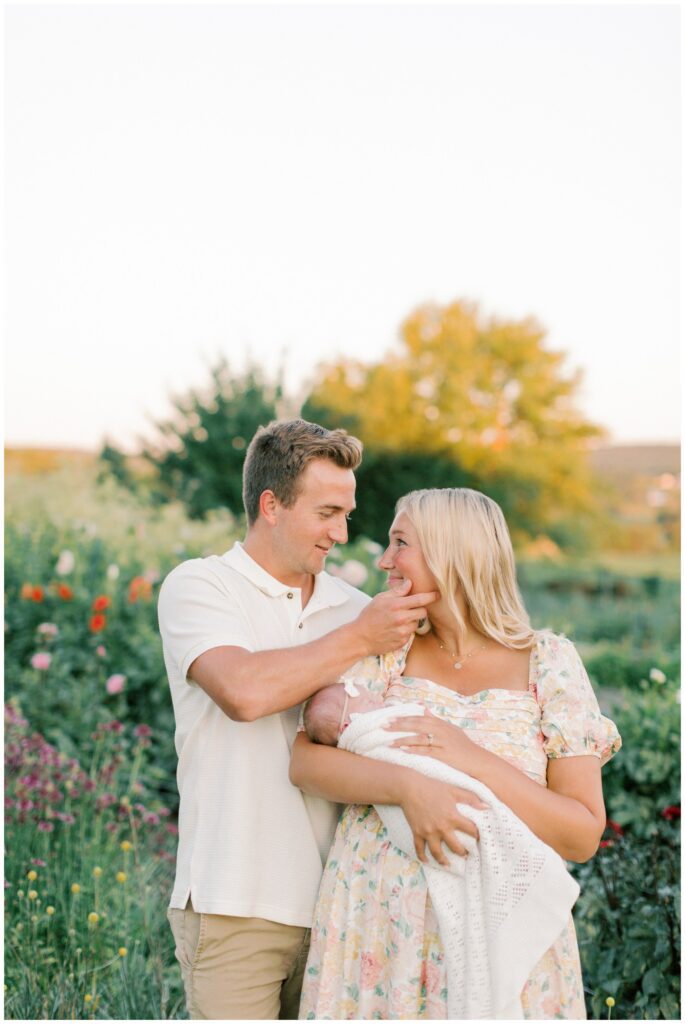 Father reaching to touch mother's face while she holds their newborn baby girl in a flower garden