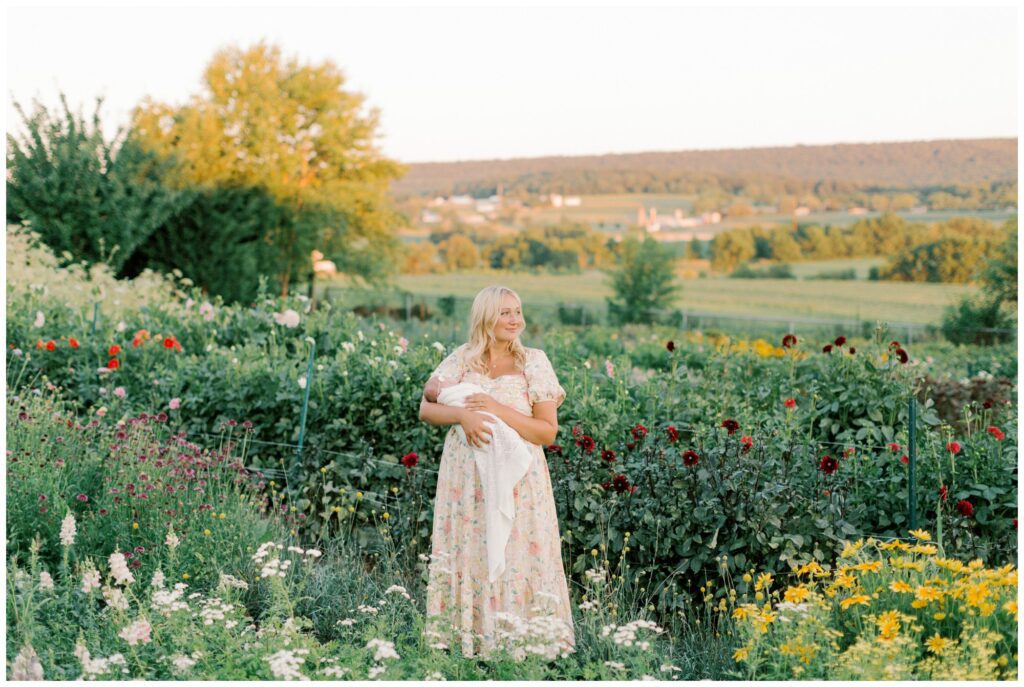 Mother looking into the distance in a flower garden while holding her newborn baby girl in a flower garden session in Lancaster, PA with Angelique Jasmin Photography